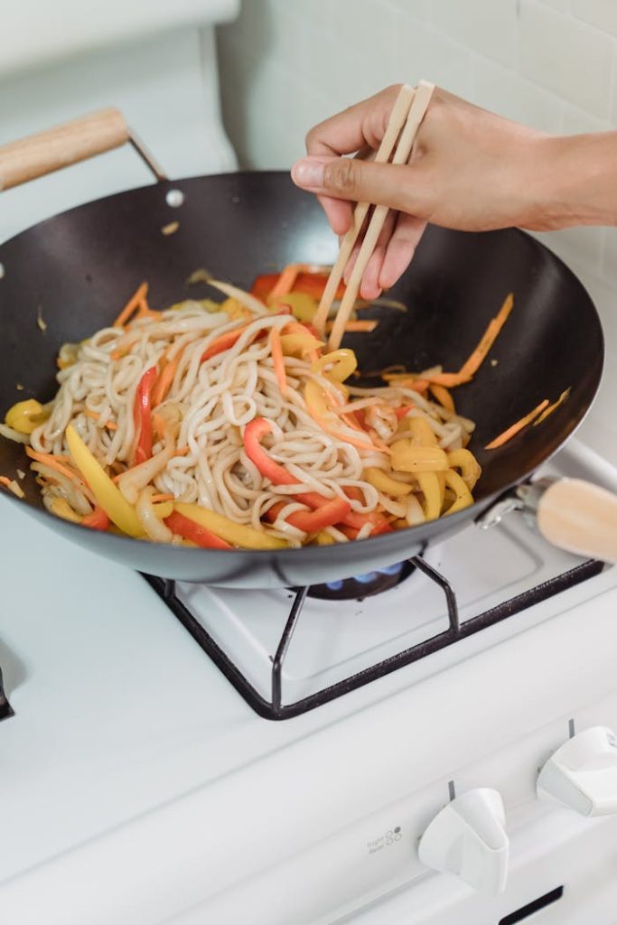 Hand using chopsticks to stir fry noodles and bell peppers in a wok on a stove.